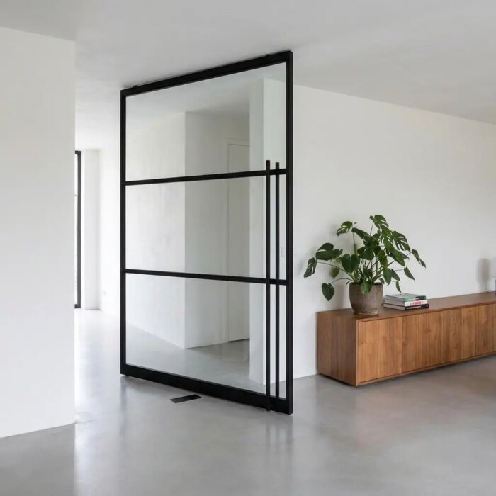 Black steel pivot door with glass standing half open in a living room with a pale grey poured concrete floor Black steel pivot door with glass standing half open in a living room with a pale grey poured concrete floor