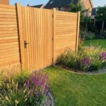 Teak gate with 50 mm slats in a teak fence behind a border of ornamental grasses